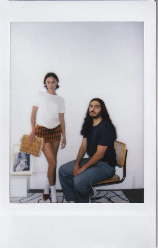 A woman stands beside a seated man in a minimalist room with white walls and a patterned rug, holding a wicker handbag. There are framed pictures on the floor next to her.