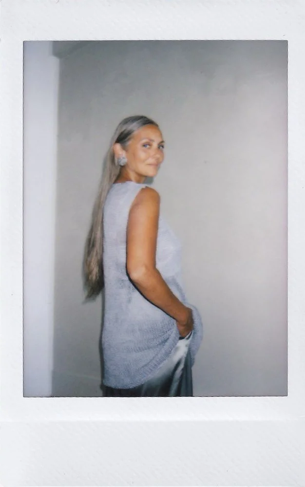 A woman with long gray hair wearing a sleeveless light gray sweater and satin pants, standing sideways with her right hand in her pocket, smiling and looking at the camera.