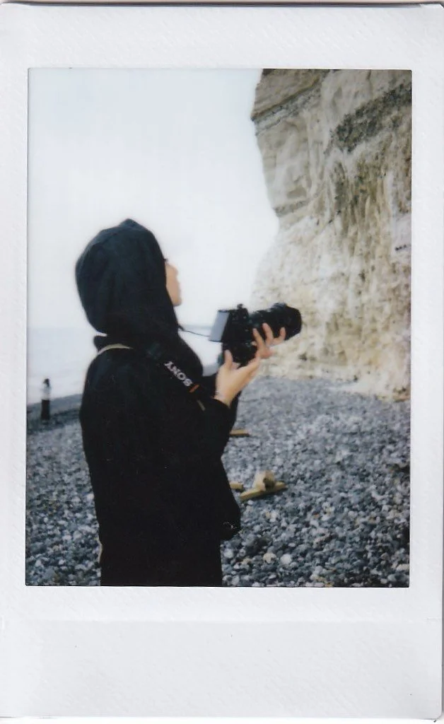Person in black hoodie holding a camera on a rocky beach with cliffs in the background.