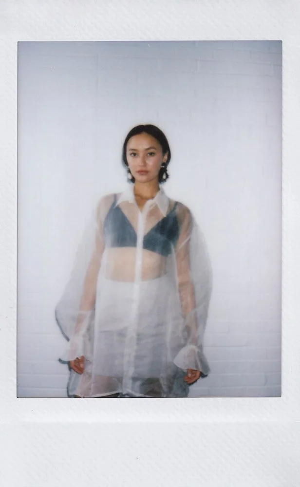 Young woman standing against a white brick wall, wearing a sheer white long-sleeve shirt layered over a dark bralette, with short black hair and earrings.