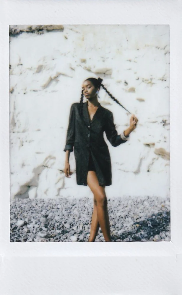 A woman with braided hair posing outdoors against a rocky, white wall, wearing a black button-up dress and holding one of her braids.