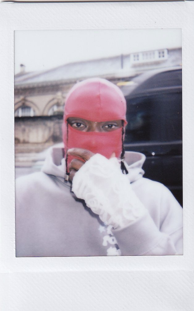 Person wearing a pink ski mask and white gloves, covering their mouth and nose, with only their eyes visible, standing outdoors in front of a building.