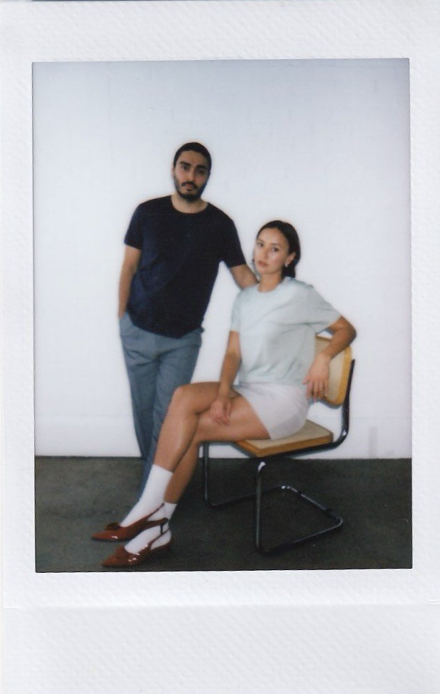 A man and woman pose in front of a plain white wall, with the woman sitting on a chair and the man standing next to her, one arm resting on her shoulder.