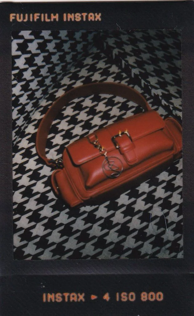 A print advertisement for Fujifilm Instax featuring a brown handbag with a top handle, embroidered logo, and buckle, placed on a black and white houndstooth patterned background.