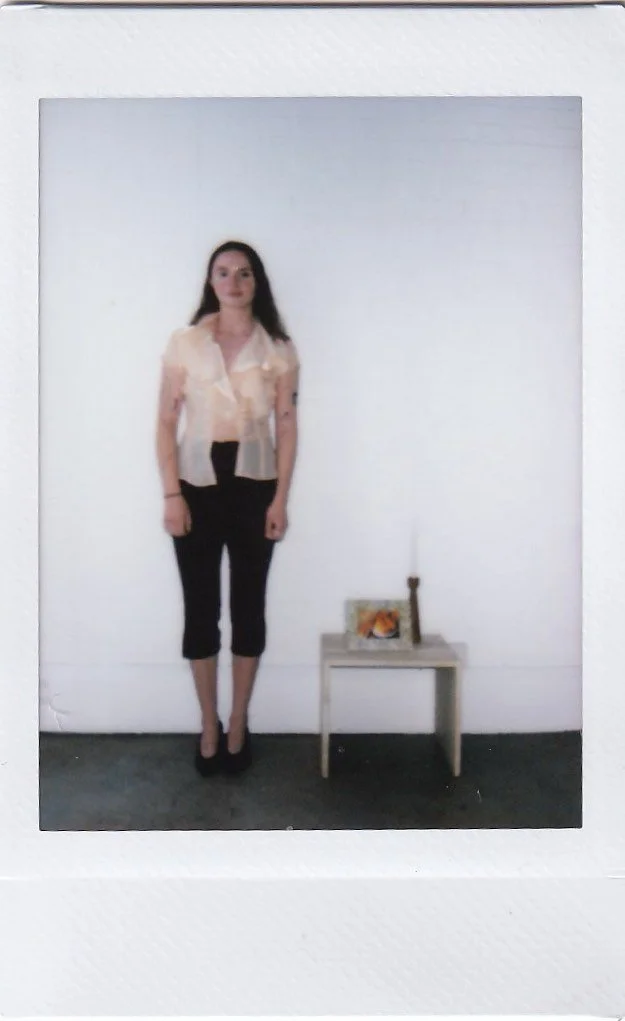 A woman with long dark hair standing against a plain white wall, wearing a beige blouse, black pants, and black heels. There is a small table with a framed photo, a candle, and a small decorative item next to her on the right.
