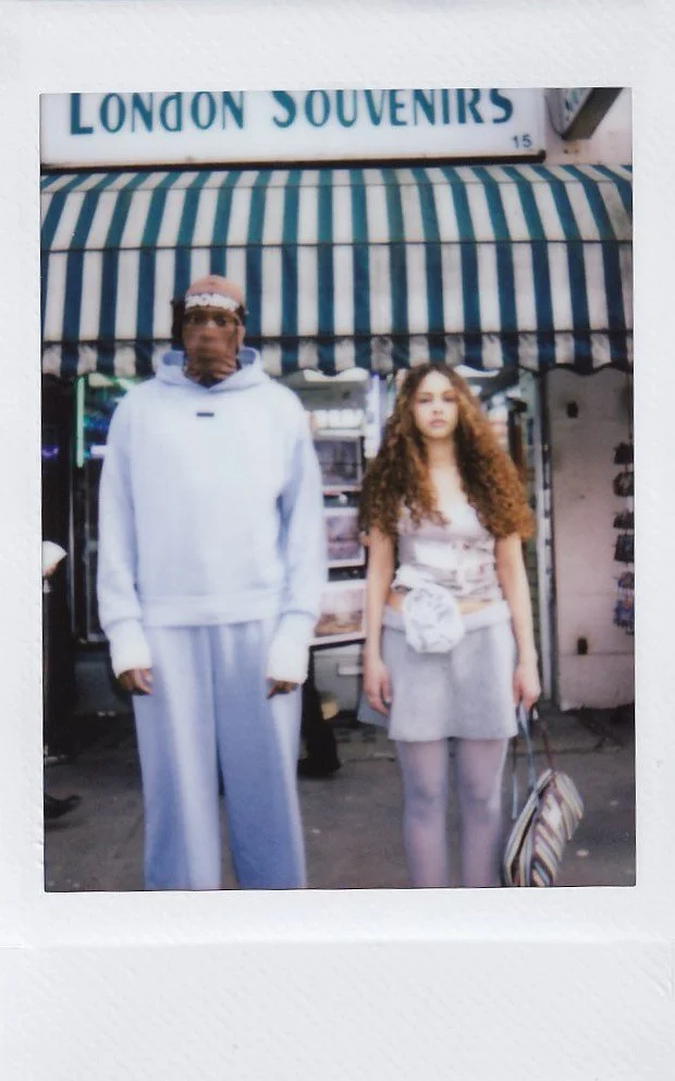 Two young women standing side by side in front of a storefront with a striped awning. One woman has curly hair and is holding a striped bag; the other woman is wearing a white hoodie and sweatpants, with a headband.