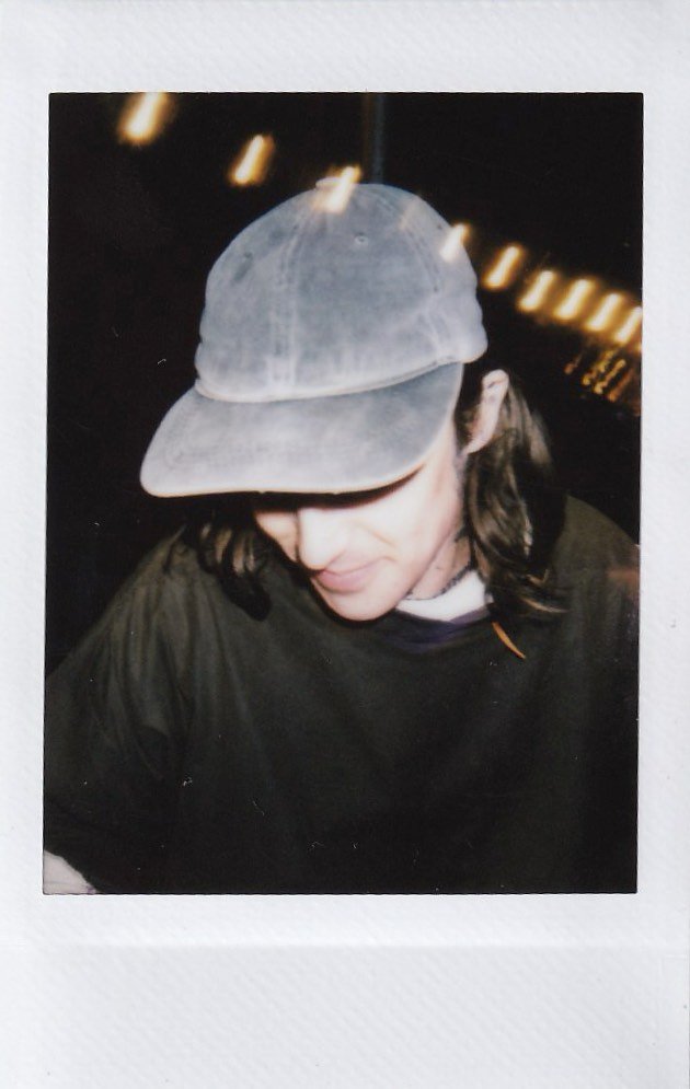 A person with long dark hair wearing a gray baseball cap and dark jacket, looking down and smiling slightly, with blurred lights in the background.