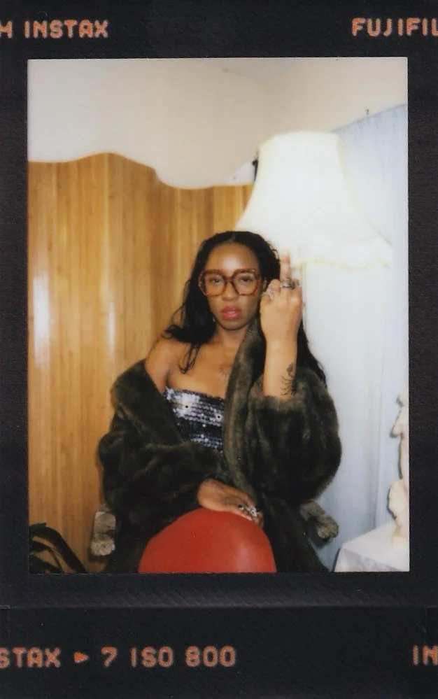 A woman sitting in a room with wood paneling behind her, wearing glasses, a fur coat, and a sparkly top, resting her hand on a large red ball.