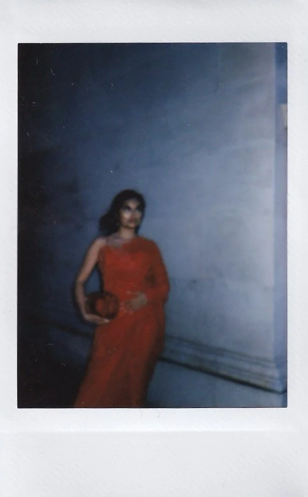 A woman in a red dress holding a clutch bag, standing against a plain wall.