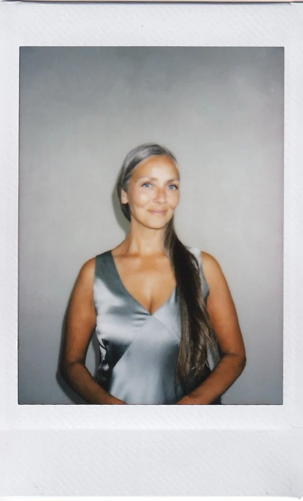 A woman with long, gray and brown hair, smiling, wearing a shiny gray and white satin sleeveless dress, standing against a gray background.
