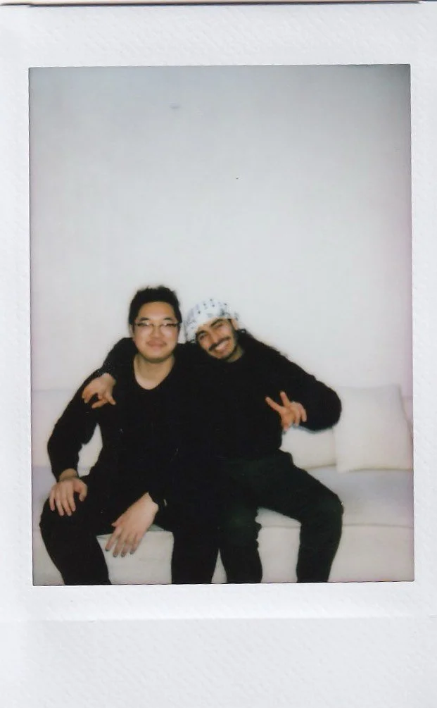 Two men sitting on a white couch against a plain white wall, with one making a peace sign and both smiling.