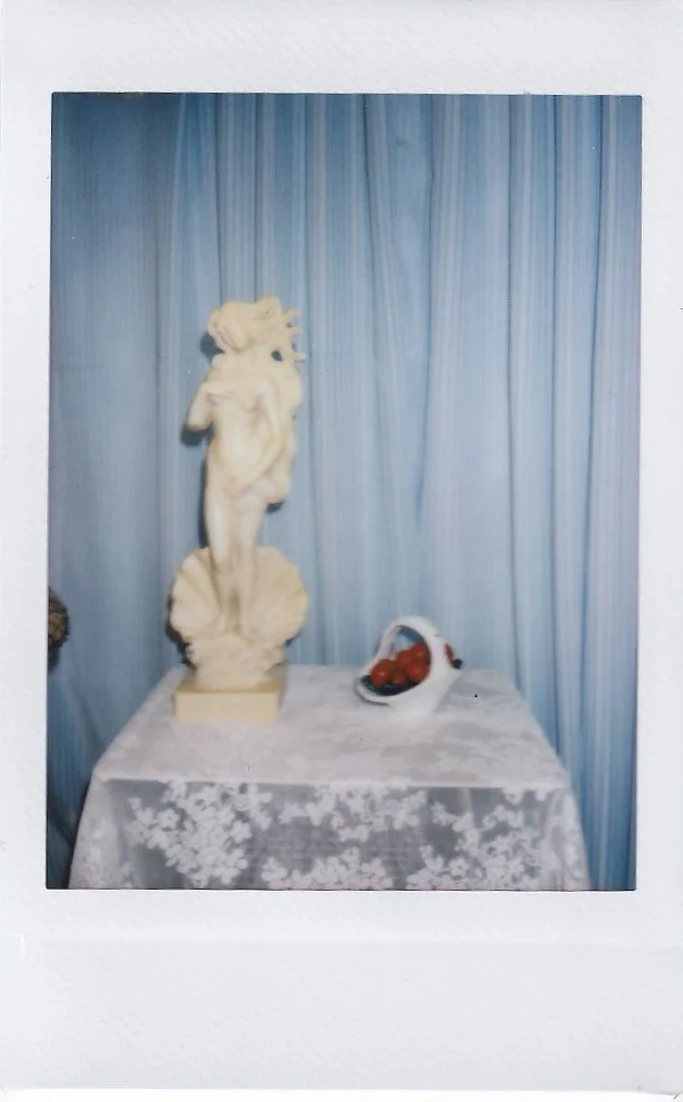 A white marble statue of a woman standing on a decorated pedestal, placed on a table covered with lace, with a blue curtain backdrop and a bowl of strawberries beside it.