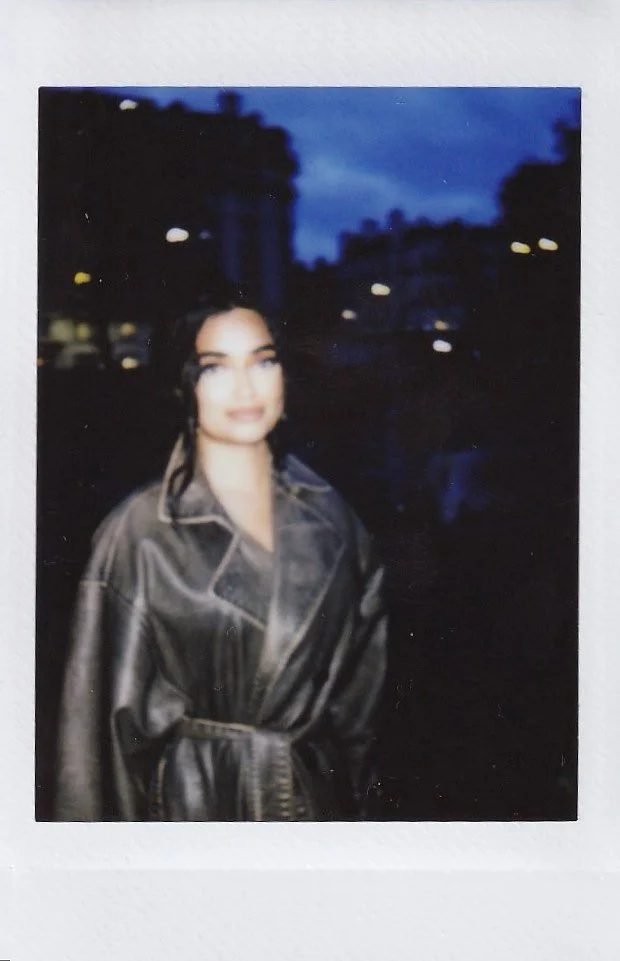 A woman in a dark leather jacket standing outdoors during dusk with illuminated streetlights and blurred buildings in the background.
