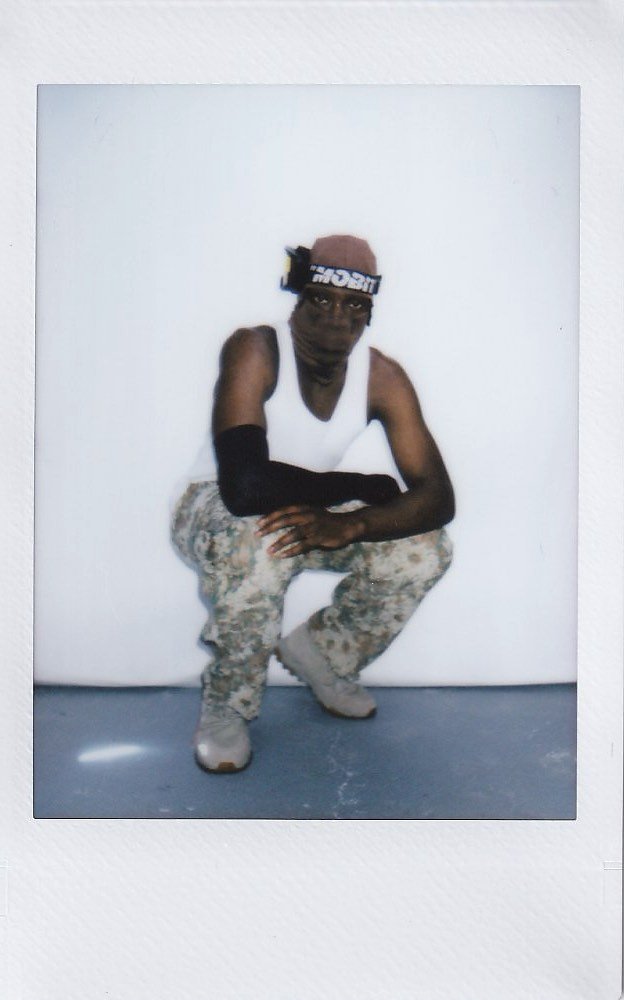Person crouching against a plain white background, wearing a brown beanie, white tank top, camouflage pants, beige sneakers, black arm sleeve, and a headband.