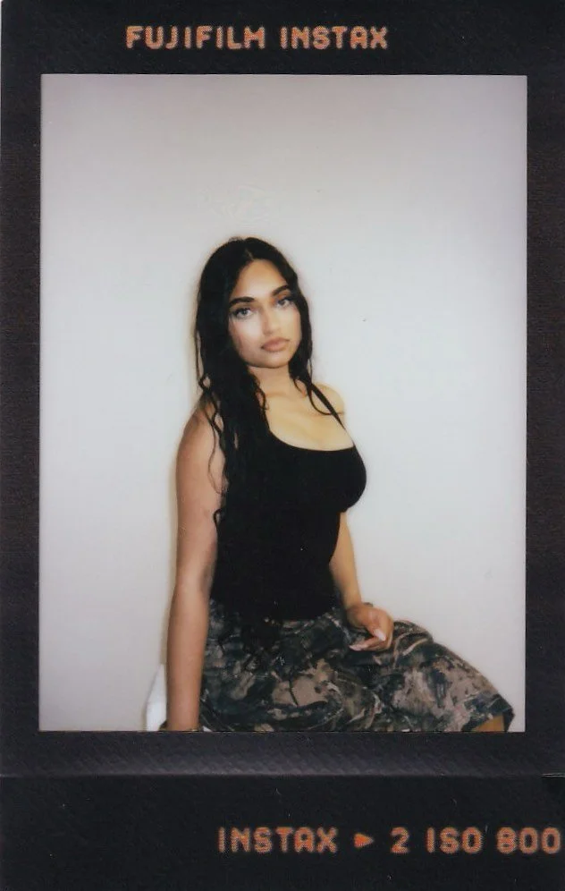A young woman with long dark hair, wearing a black tank top and camouflage pants, sitting against a white wall with a black photo border, with the words 'Fujifilm Instax' above and 'Instax   2 150 800' below.