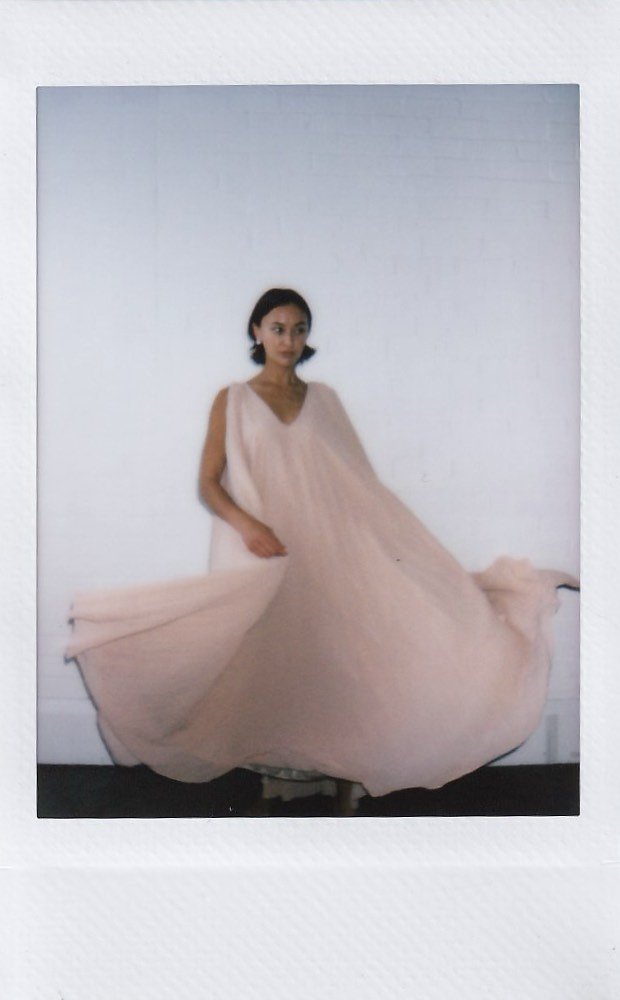 A woman in a flowing, pale pink dress standing against a plain white wall.