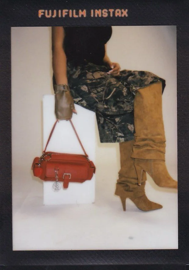 A person sitting on a white block, wearing a black top, camouflage pants, and tan knee-high boots. They hold bag with a strap in their left hand, which rests on the white block. The background is plain, and the photo is branded with 'Fujifilm Instax'