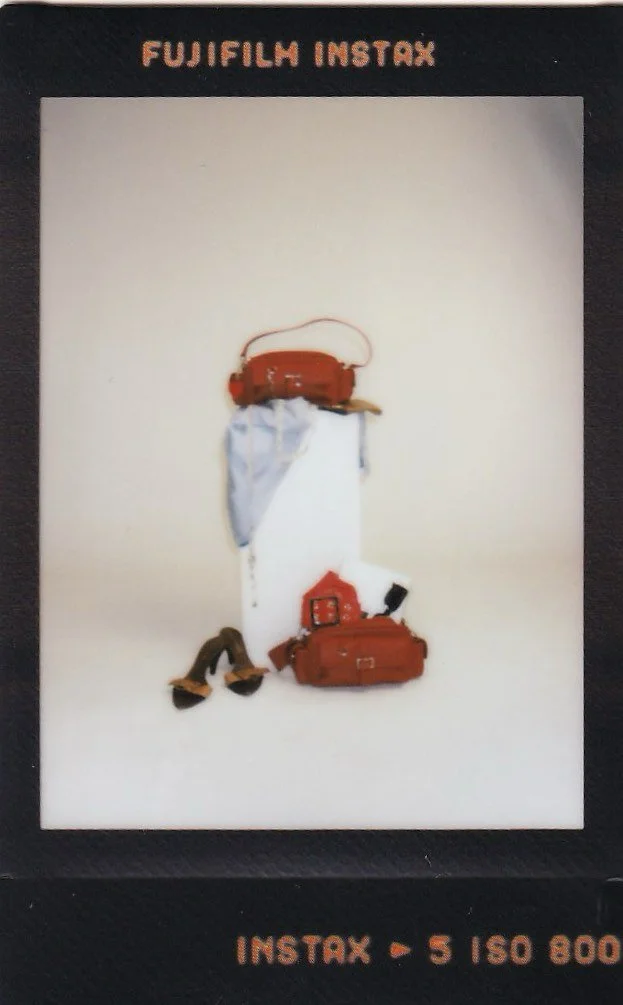 Fujifilm Instax photo of a travel outfit including a brown hat, white shirt, brown sandals, a small red house-shaped purse, a brown bag, and travel accessories, with the text 'INSTAX   5 ISO 800' at the bottom.