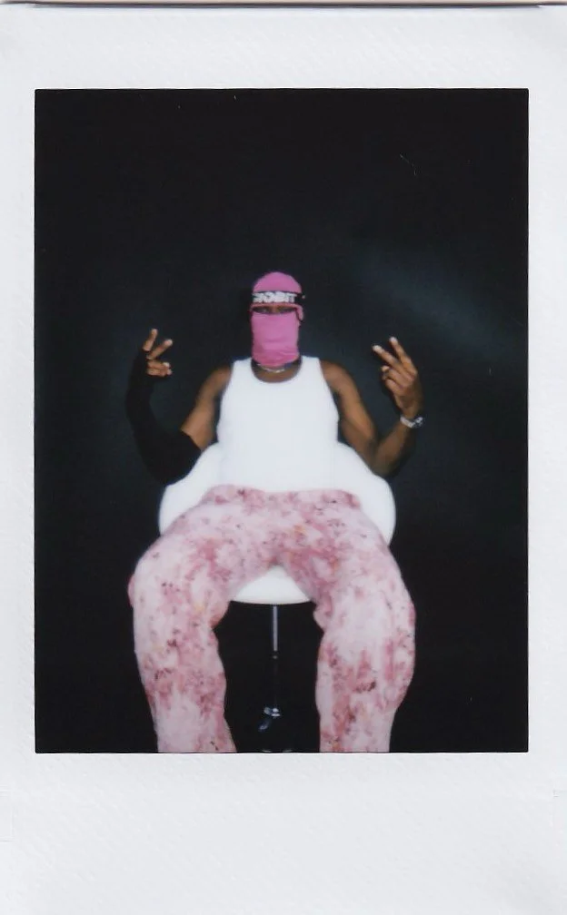 Person sitting on a chair against a black background, wearing a pink bandana covering face, a white tank top, pink tie-dye pants, and making peace signs with both hands.