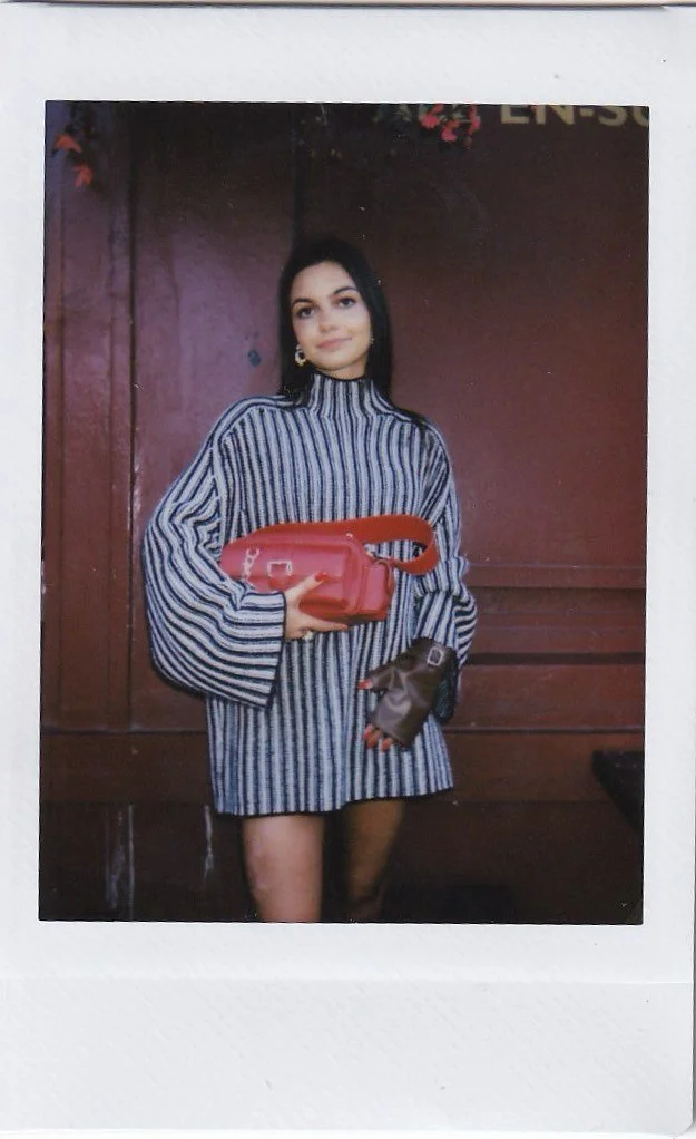 A woman standing in front of a wooden wall, holding a small orange bag with a handle and a wallet or phone in her other hand, wearing a striped long-sleeve dress with a high neckline.