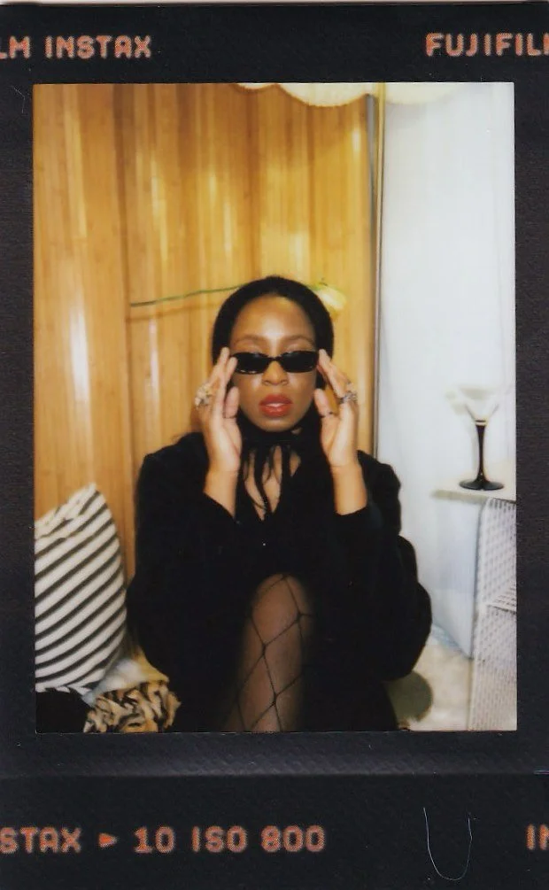 A woman with dark hair and sunglasses adjusting her glasses, sitting in a room with wooden paneling and white curtains, wearing a black outfit and fishnet stockings.