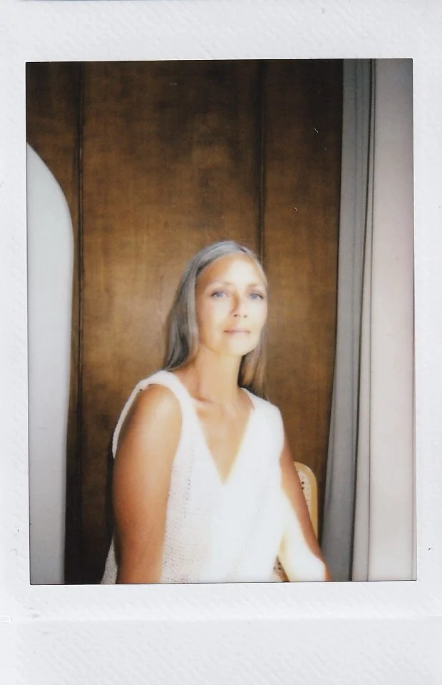 A woman with gray hair and light skin sits in front of a wooden wall, wearing a sleeveless white top, looking at the camera with a soft expression.