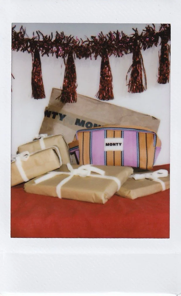 Wrapped gifts, a striped Monty bag, and a newspaper in front of a decorative garland against a white wall.