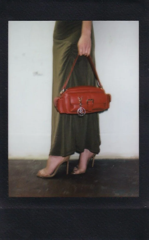 A person wearing a long green skirt and high heels, holding a red handbag with a circular keychain, standing against a plain white wall.