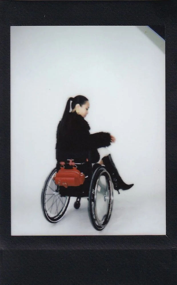 A woman in a wheelchair with a black coat and black boots, carrying a brown bag, against a white background.