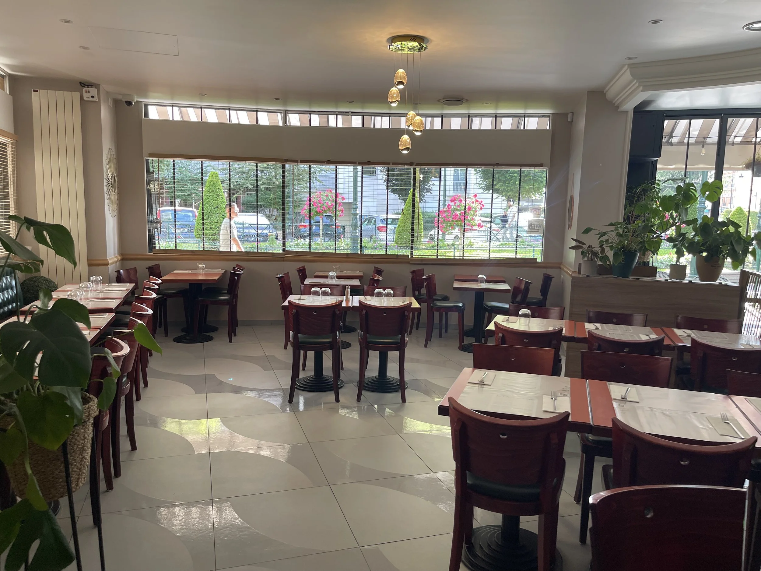 Salle à manger d'un restaurant bien éclairée, avec des tables en bois, des chaises en bois foncé, des plantes vertes et des grandes fenêtres laissant passer la lumière naturelle.