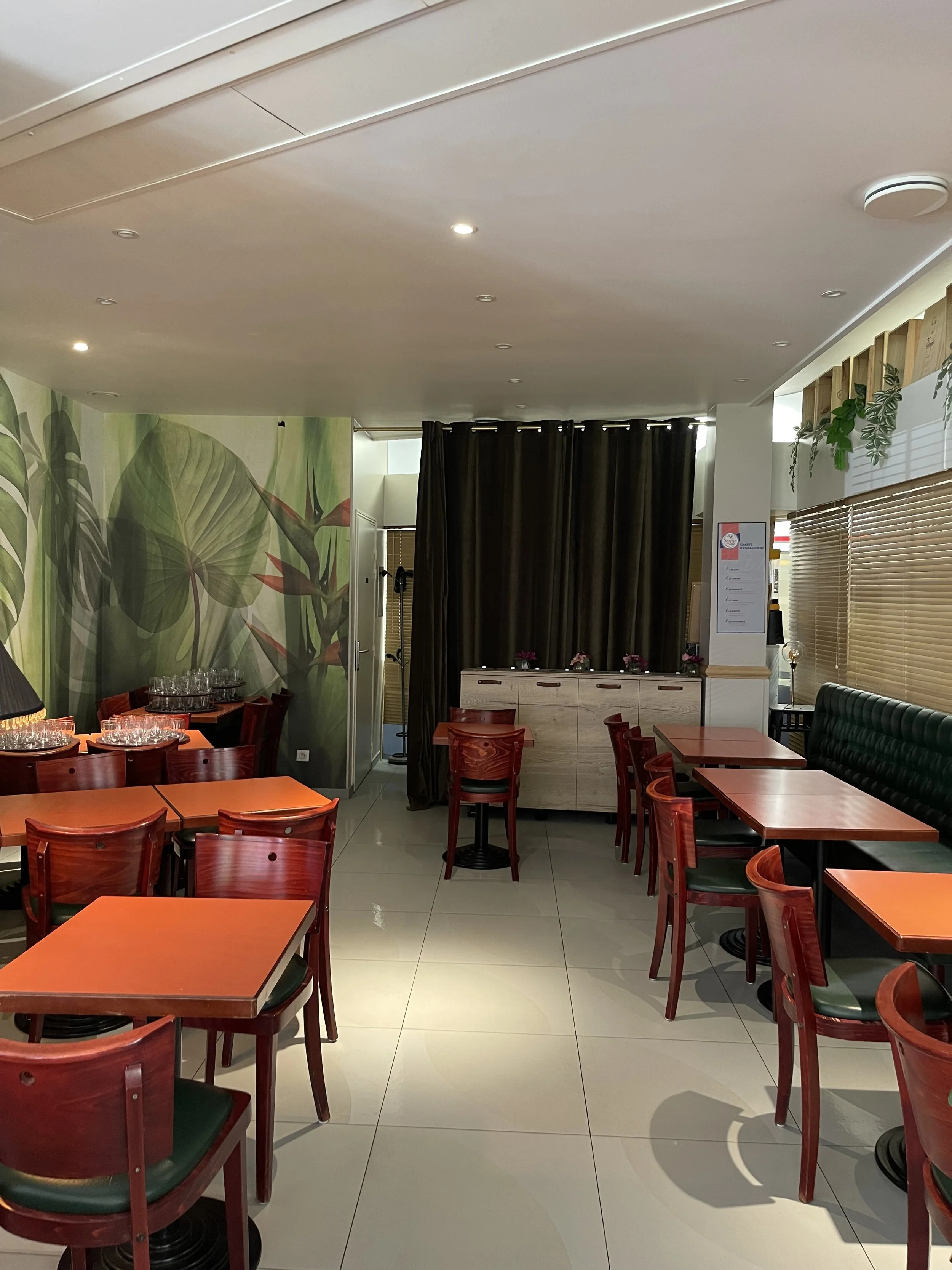 Salle de restaurant avec tables en orange, chaises en bois rouge et un mur avec une grande fresque de feuilles vertes.