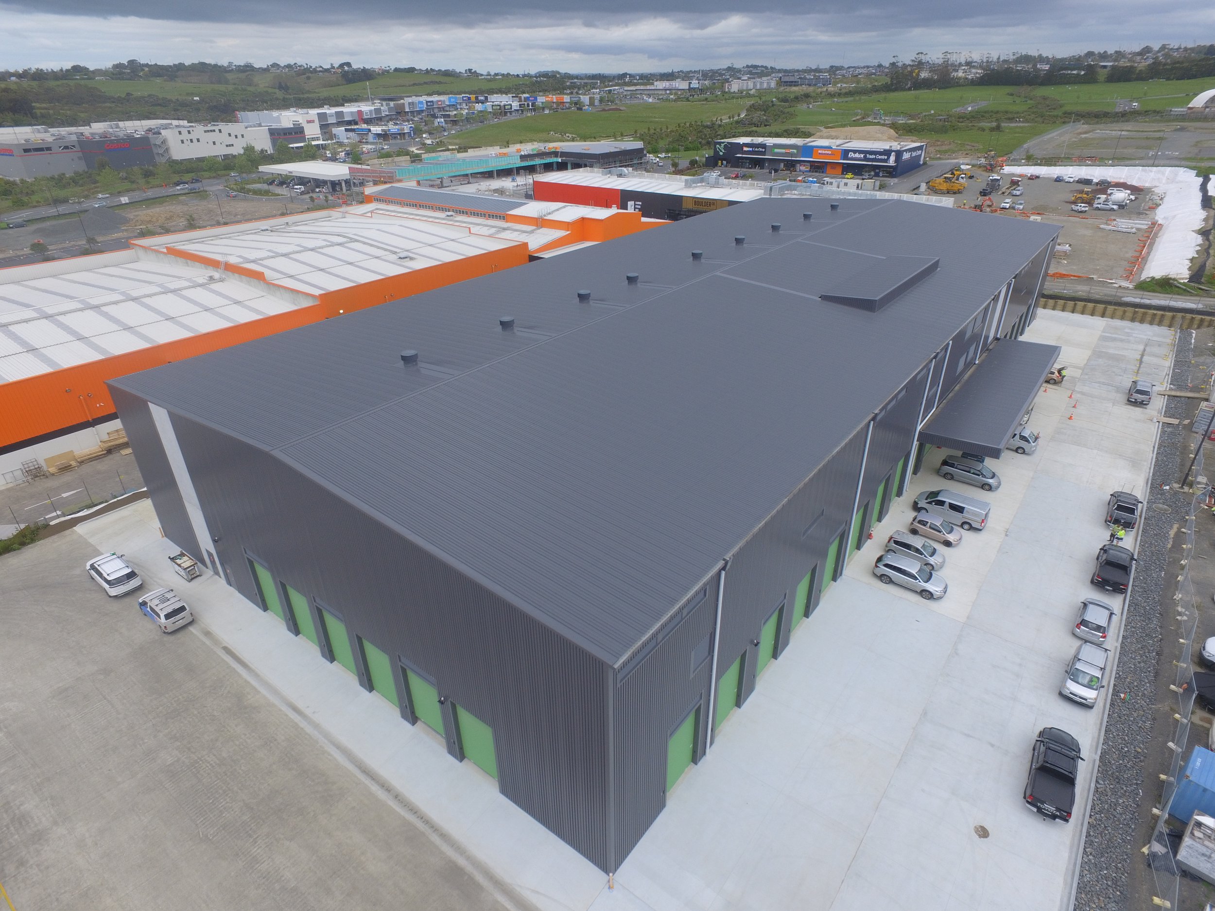 Aerial view of a large modern warehouse with a black metal roof, green loading bays, surrounded by a parking lot with cars, in an industrial area with other warehouses and green fields in the background.