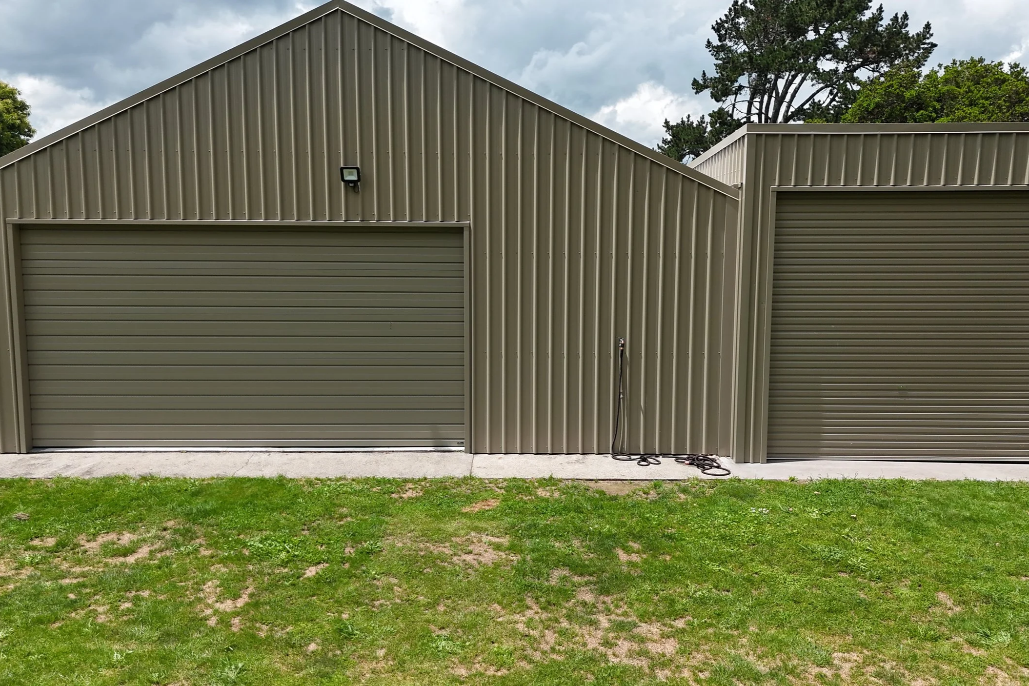 Two tan metal garages with roll-up doors, one double and one single, situated on a concrete pad with a grassy lawn in the foreground, under a cloudy sky, with some trees in the background.