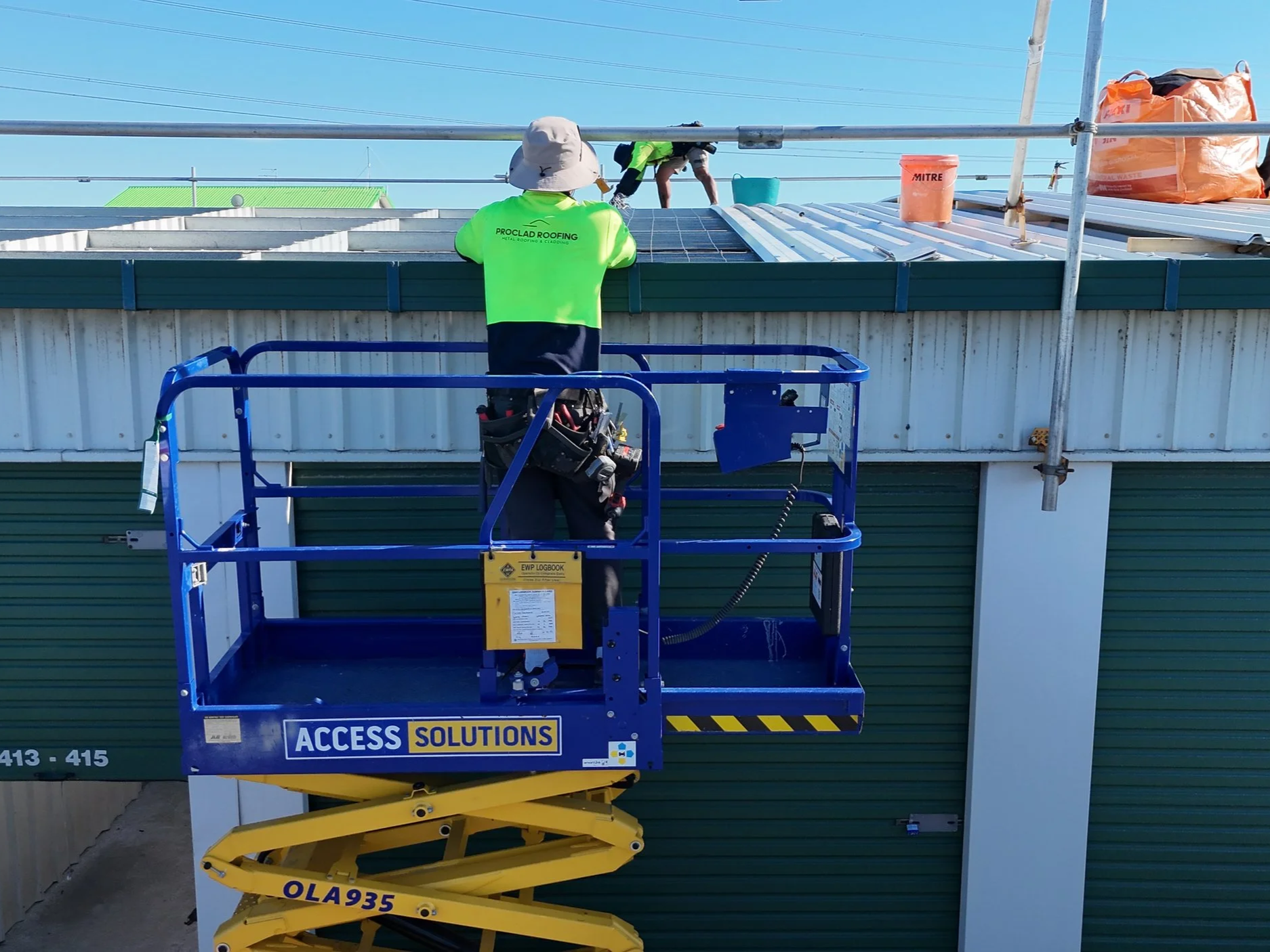 A worker on a blue lift working on a metal roof with tools and supplies nearby at a construction site.
