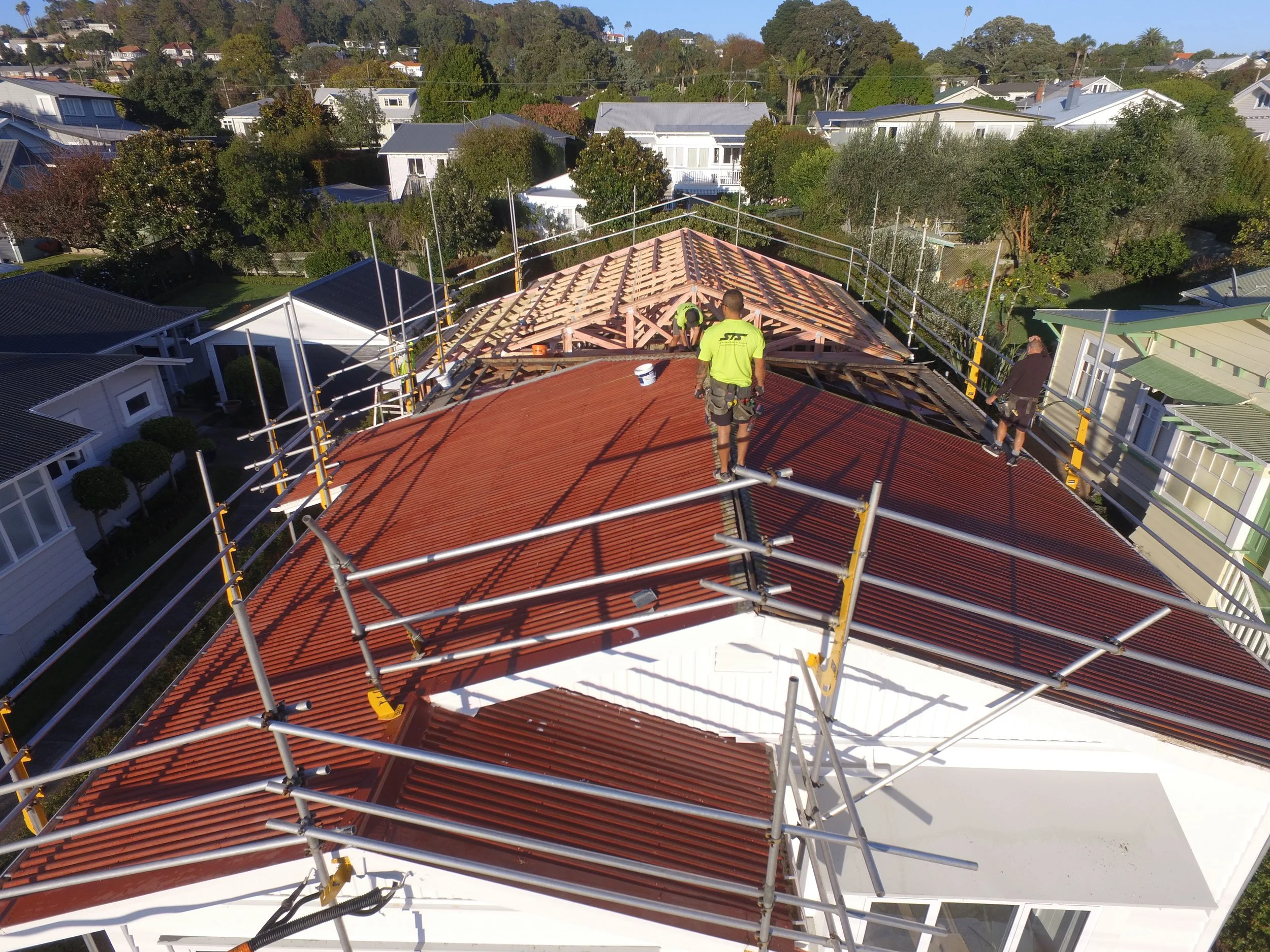 Construction workers installing new red metal roofing on a house, surrounded by scaffolding, in a residential neighborhood.