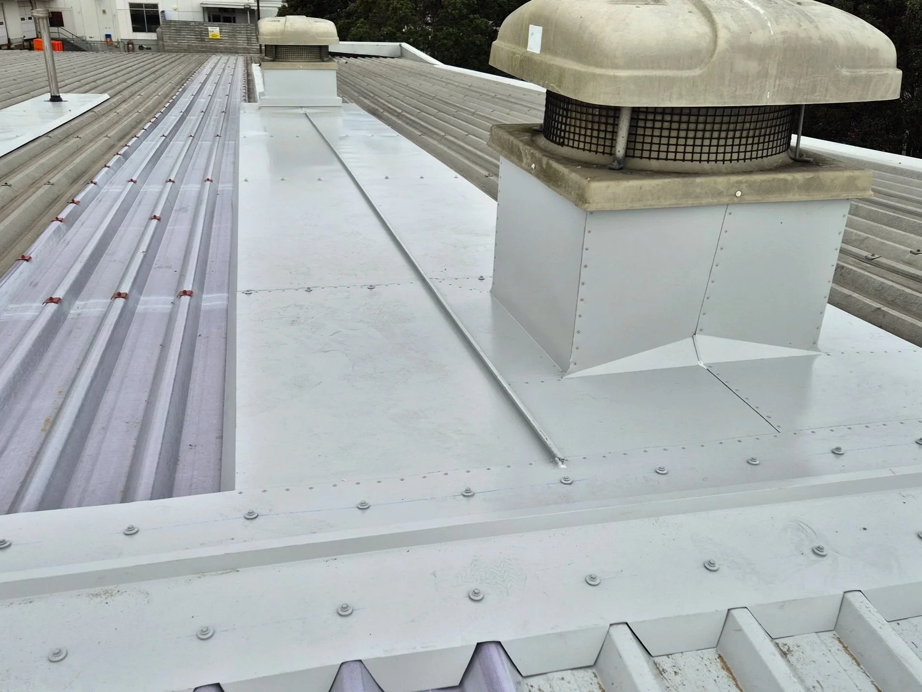 View of a metal rooftop with two large ventilation units installed on it, featuring metal and concrete materials, with a background of trees and a building.