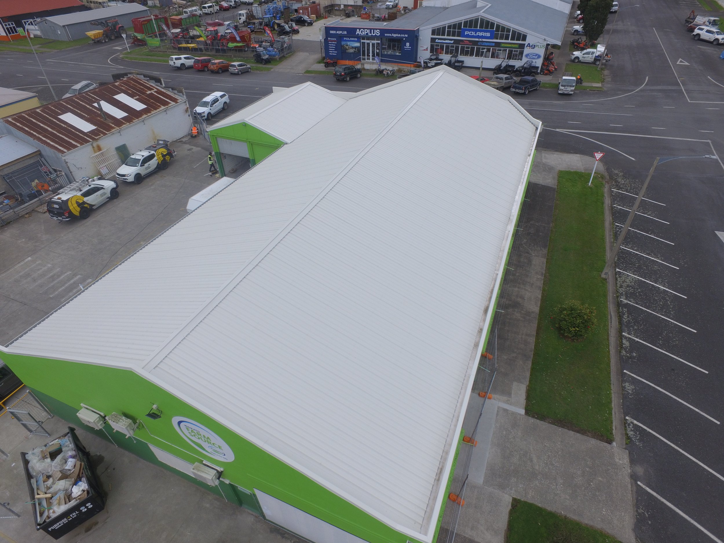 Aerial view of a commercial building with a white metal roof, green walls, and parking lot surrounding it, next to other shopfronts and parked cars.