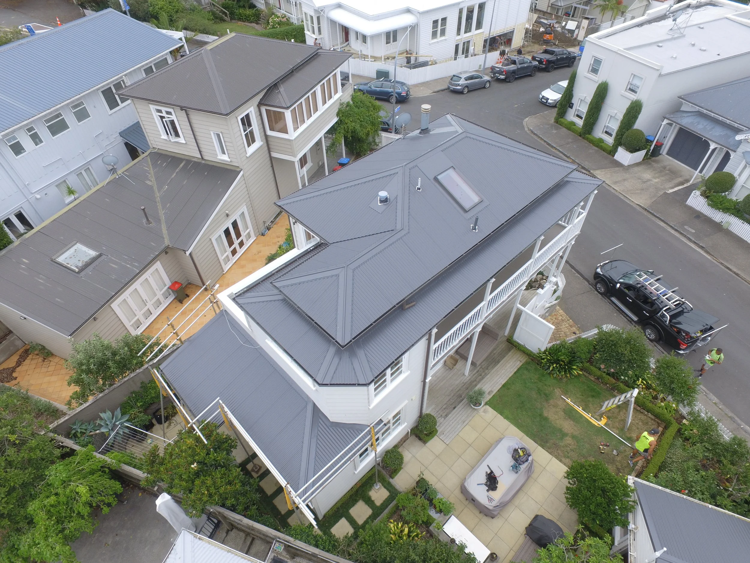 Aerial view of a neighborhood with multiple houses, focusing on a large two-story house with a gray metal roof, surrounded by green trees and parked cars.