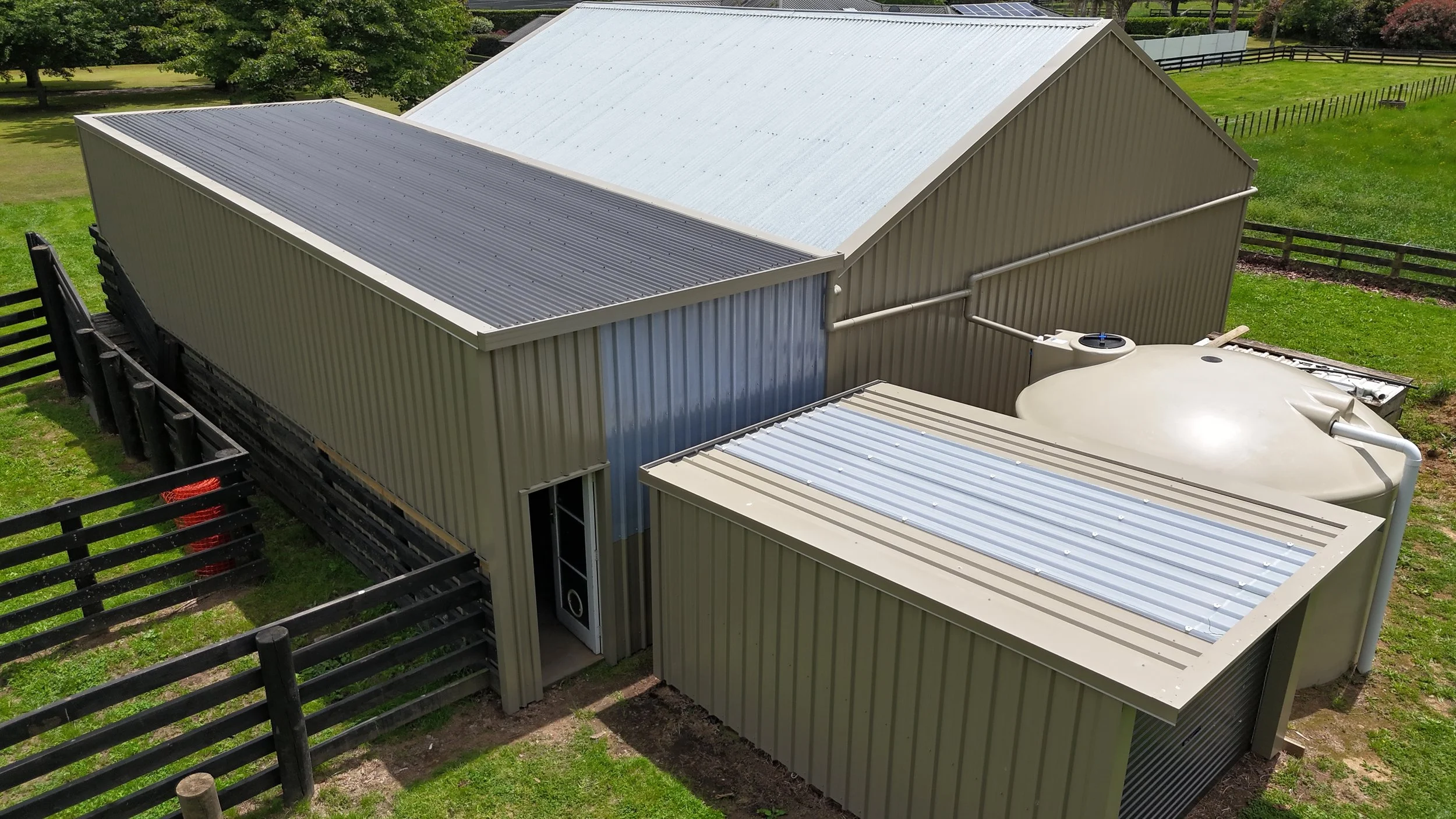 An aerial view of a metal building with two sections, one with a light gray roof and the other with a blue door, adjacent to a large white water tank, surrounded by a black wooden fence and green grass.