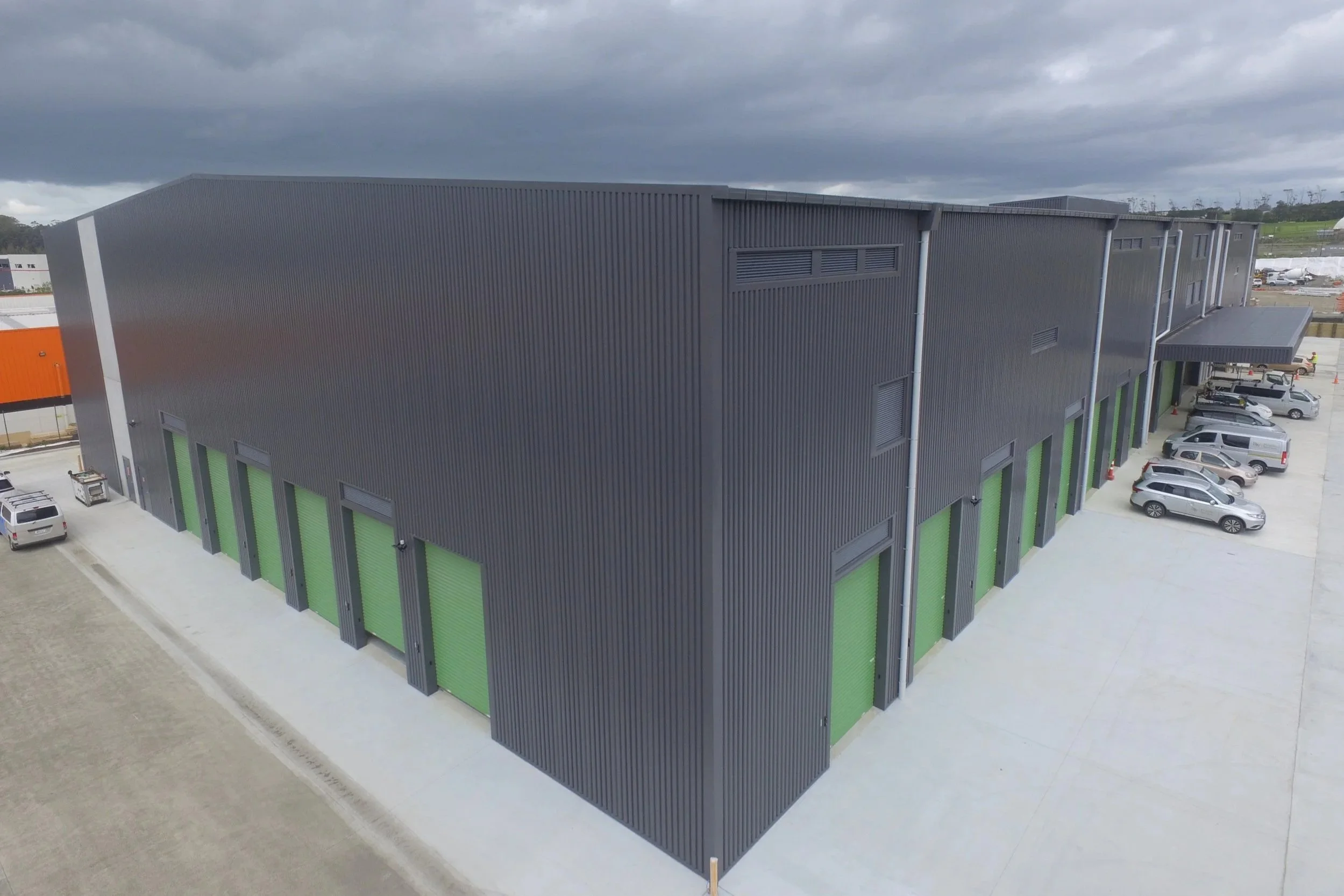 Large modern industrial building with black ribbed metal exterior and green storage unit doors, parking lot with cars, overcast sky.