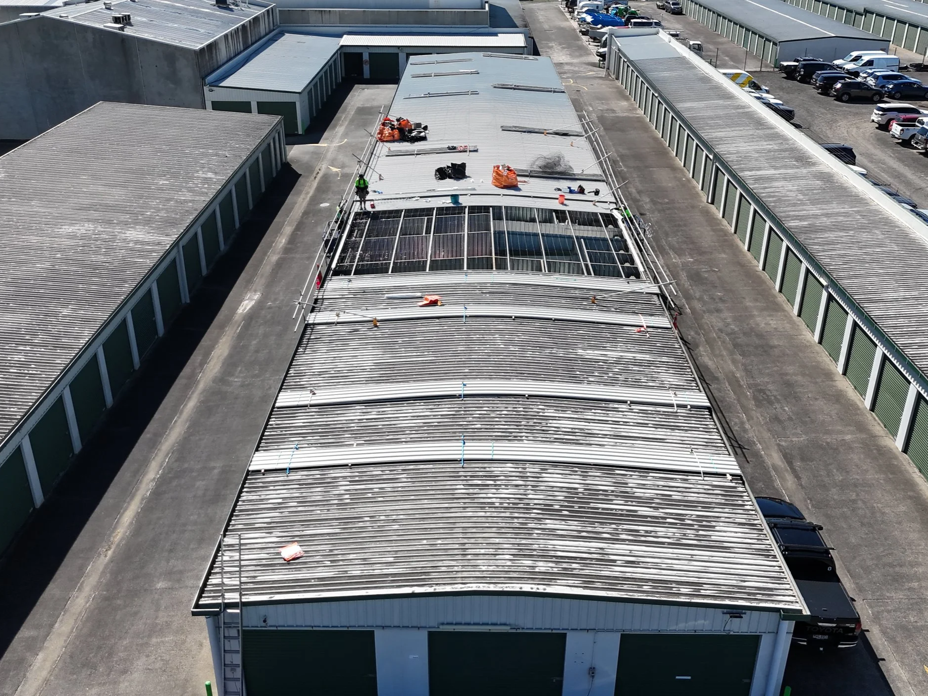Aerial view of a building with a corrugated roof undergoing maintenance or installation, with tools and equipment on the roof and workers present. Surrounding parking lot with parked cars.