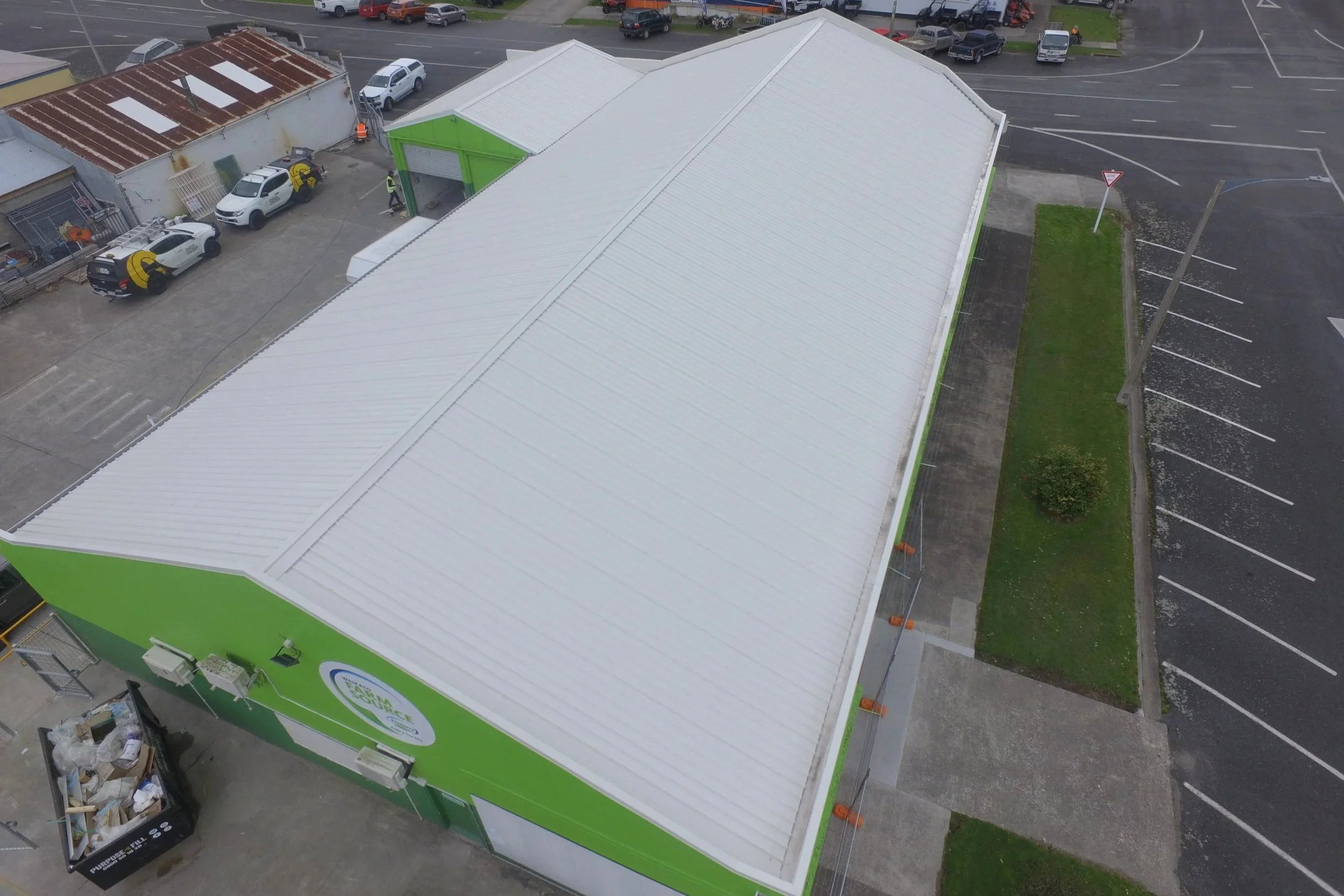 An aerial view of a large commercial building with a white metal roof, a green wall, and a parking lot with several parked cars and a designated recycling area with trash bins.