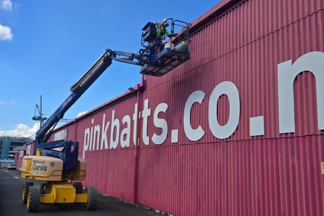 Workers on a cherry picker installing white letters on a pink corrugated metal wall that reads 'pinkbats.co.n'.