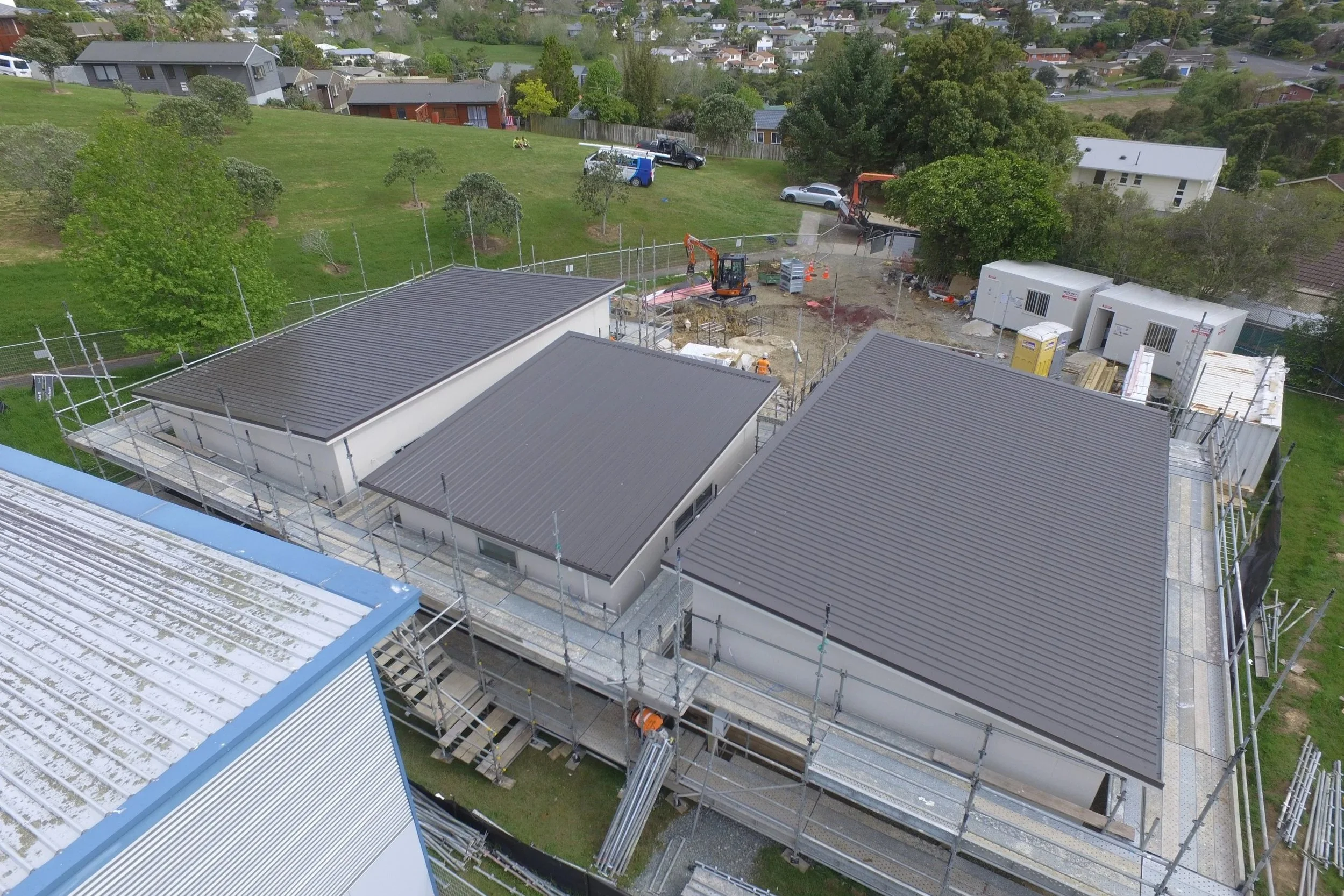 Aerial view of a construction site with three single-story buildings with gray metal roofs, scaffolding surrounding the structures, construction equipment, and materials. In the background, there are residential houses, trees, and a grassy hillside.