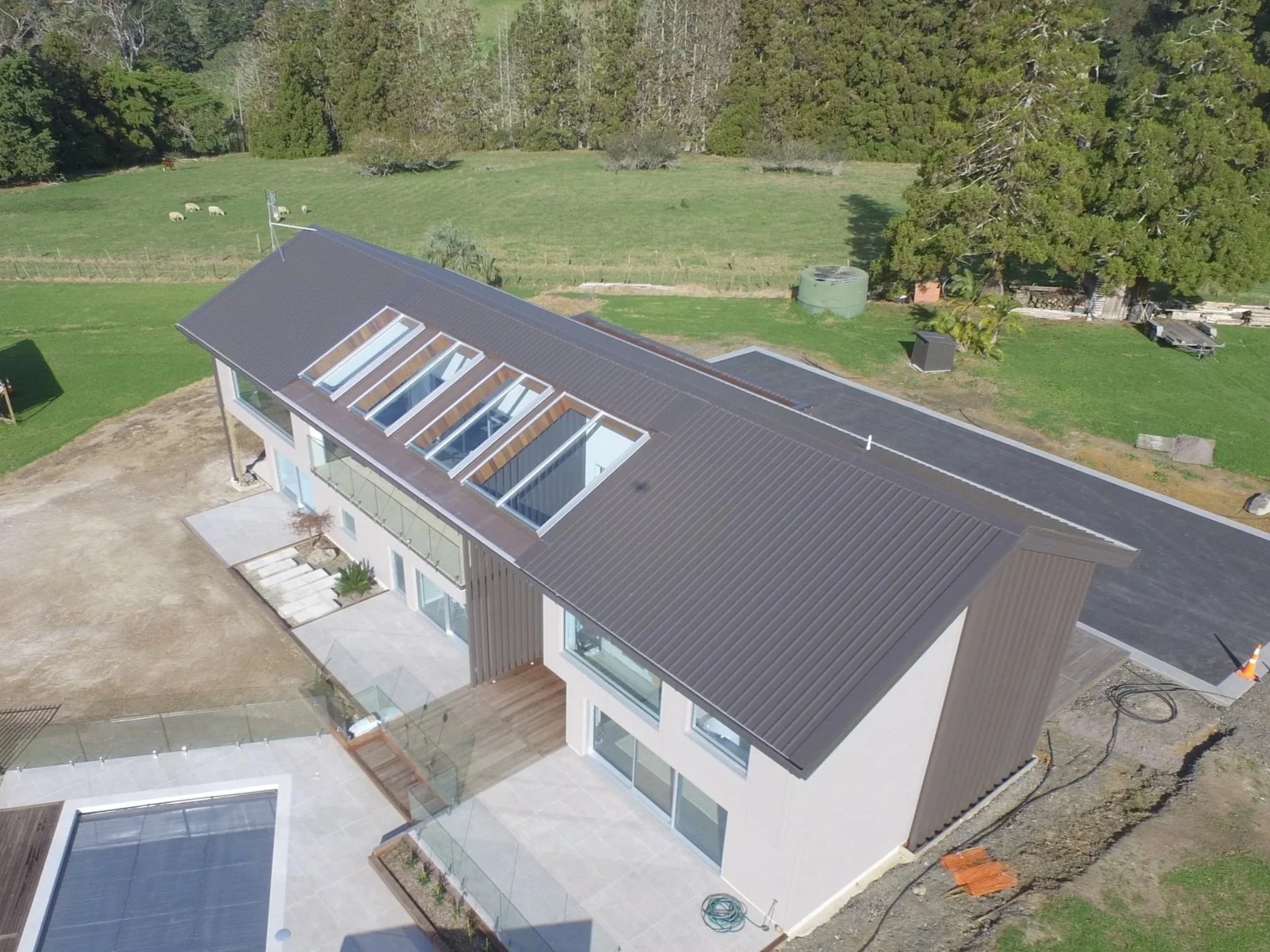 A modern two-story house with a metal roof, large windows, and a ground-level patio, situated in a rural area with green fields, trees, and grazing sheep in the background.