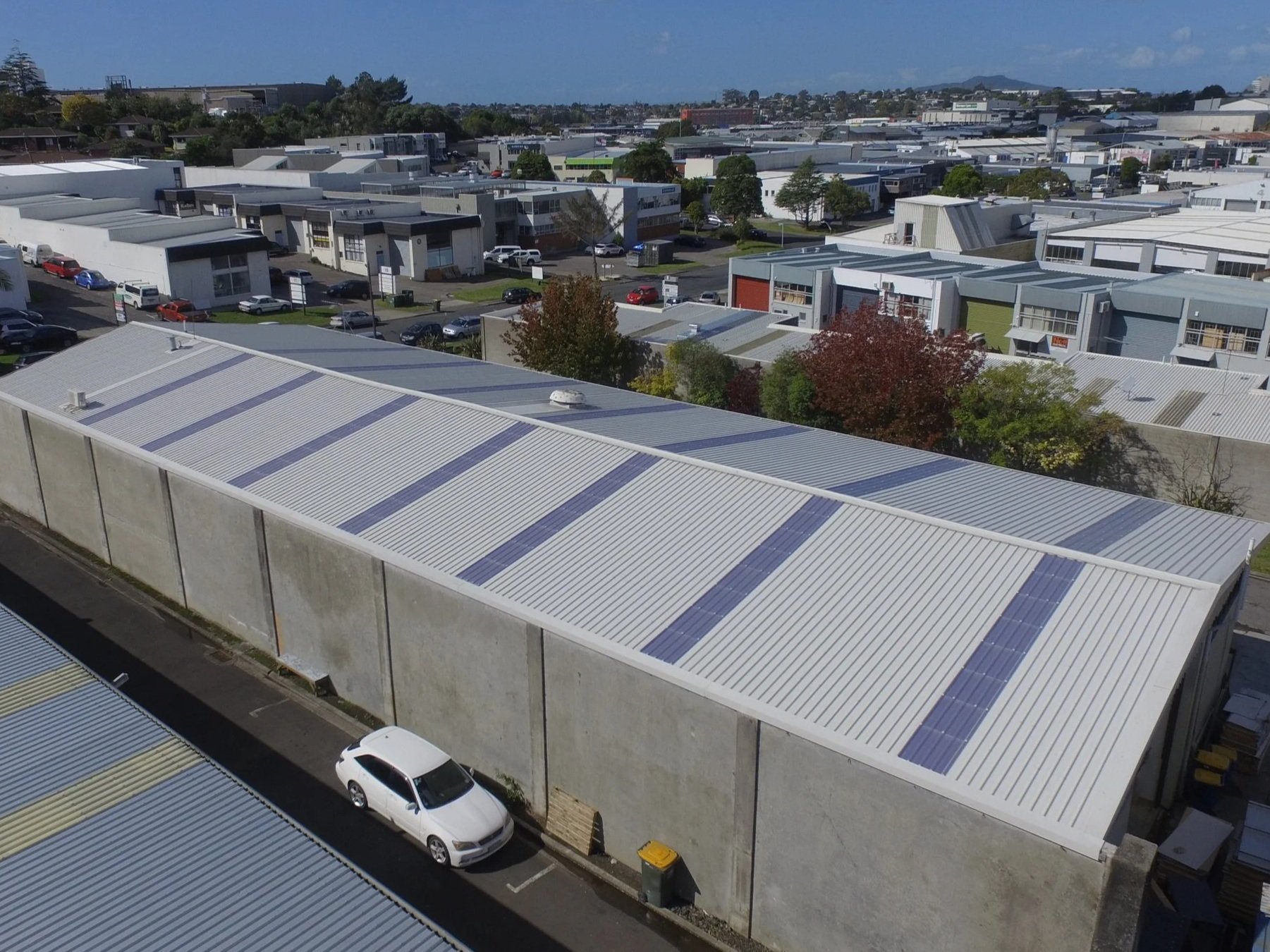 Aerial view of industrial buildings with solar panels on the metal roofs, parking lot with cars, and trees with fall foliage in a commercial area.