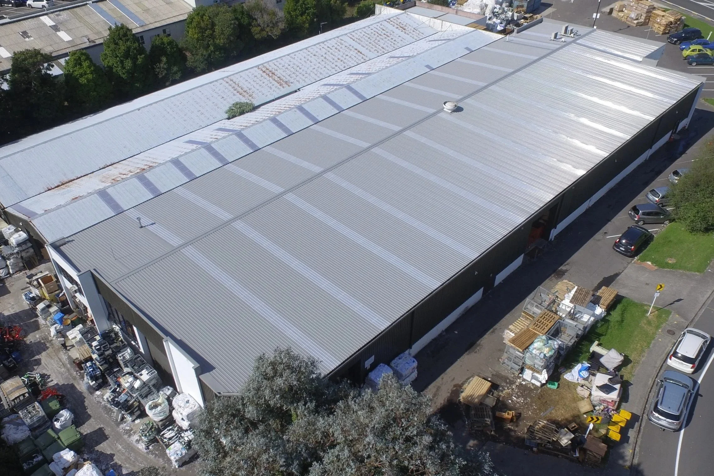 Aerial view of a large industrial warehouse with a metal roof, surrounded by parked cars, pallets, and outdoor storage areas.