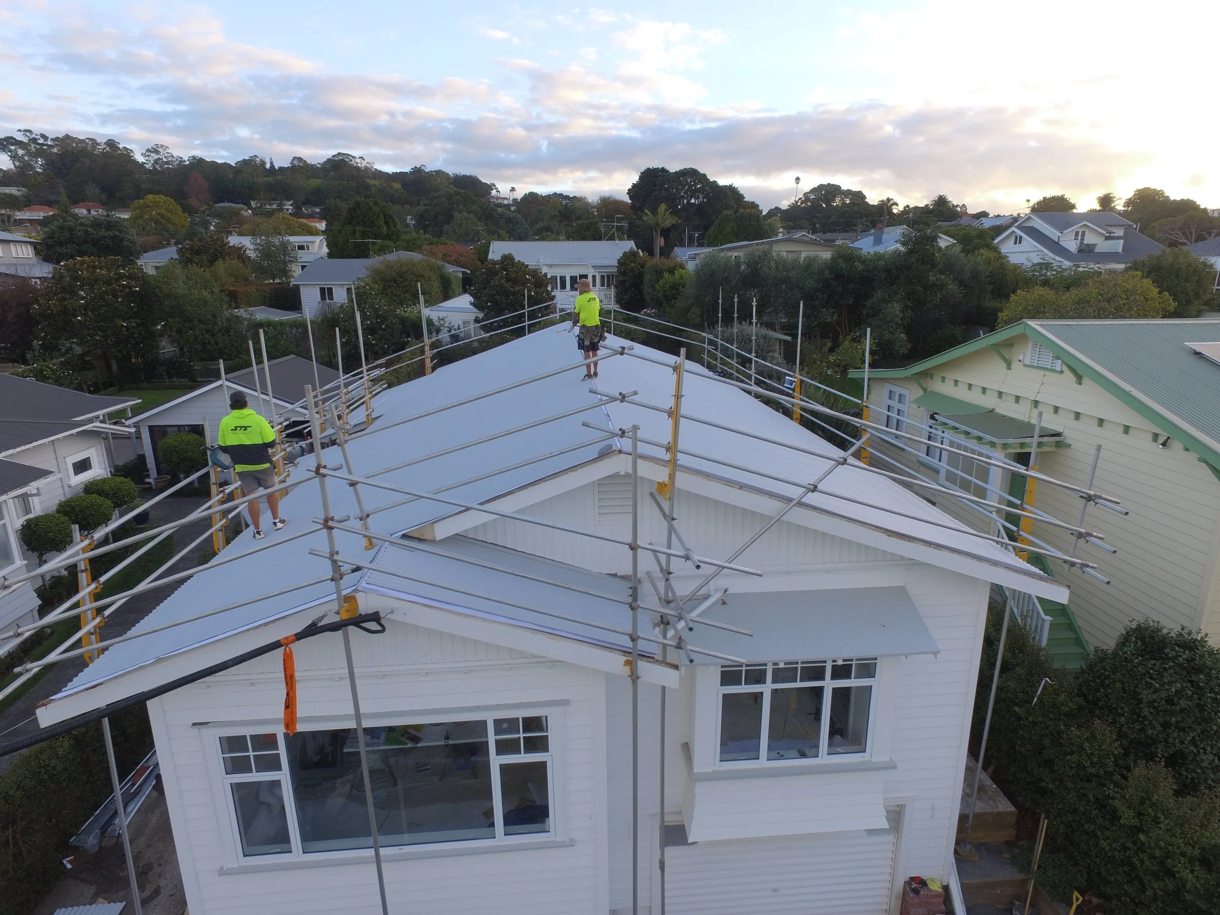 Construction workers installing or working on the roof of a white house, surrounded by scaffolding, in a residential neighborhood during daytime.