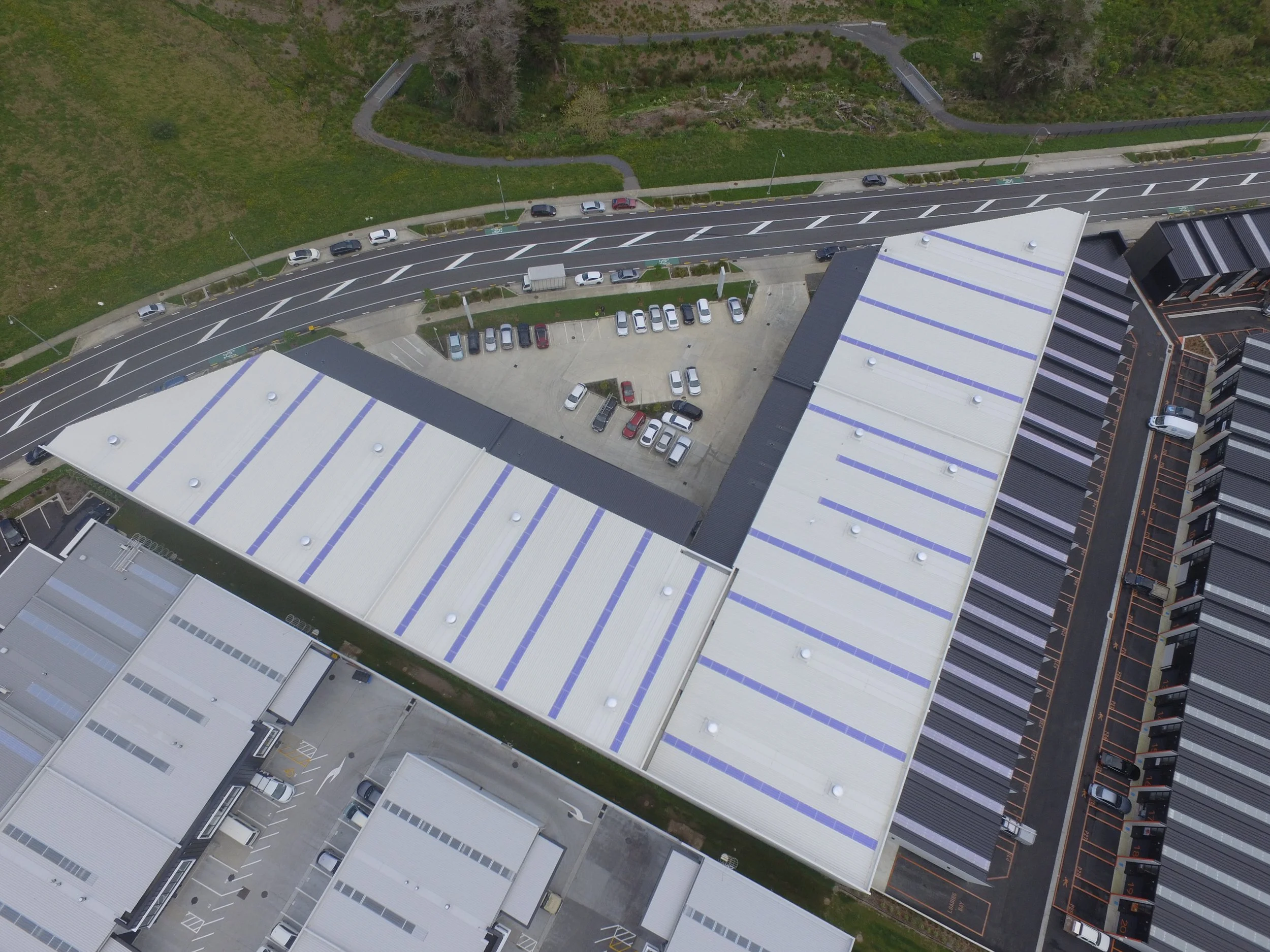 Aerial view of a modern commercial building with a white roof, adjacent parking lot, and surrounding roads, residential area, and green space.