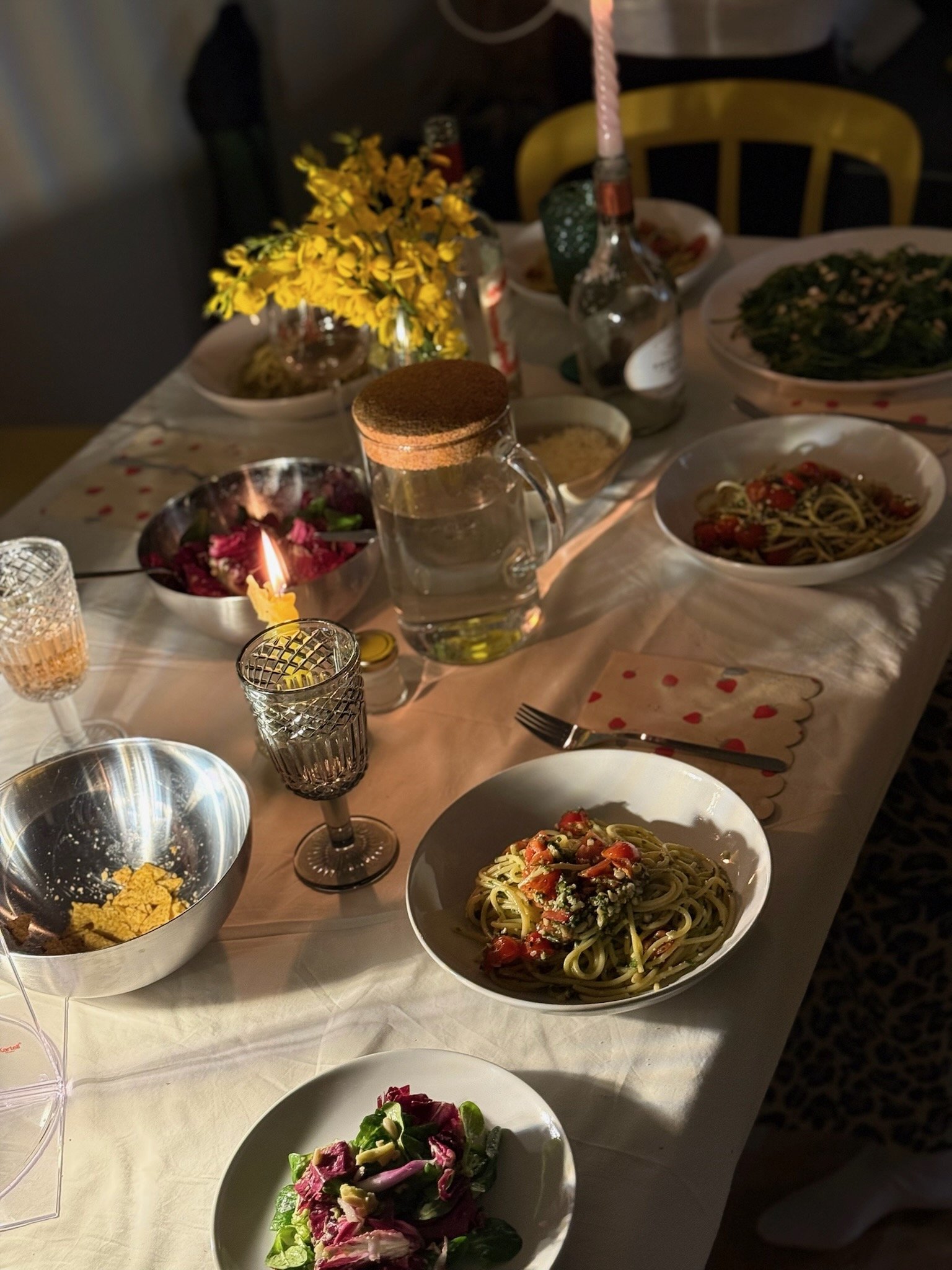 Tavola apparecchiata con piatti di pasta, insalata, fiori gialli, bicchieri di vetro, brocca d'acqua e candela accesa.